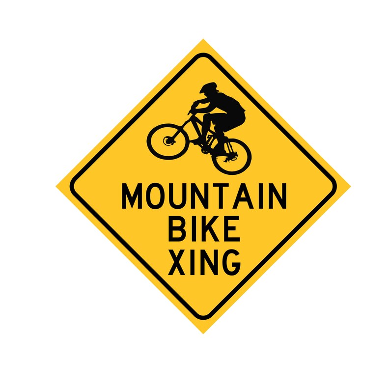 Op de afbeelding: Een geel, ruitvormig waarschuwingsbord met zwarte tekst en een silhouet van een persoon die op een mountainbike rijdt. Het bord luidt "Mountain Bike Xing".