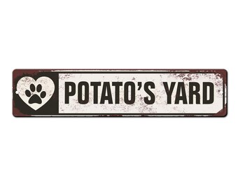 May include: A white metal sign with a brown border and distressed texture. The sign reads "POTATO'S YARD" in black text. A white heart with a black paw print is on the left side of the sign.
