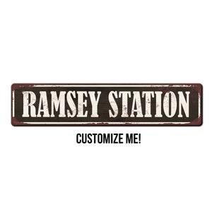 May include: Rustic metal sign with the text "RAMSEY STATION" in white lettering on a black background. The sign has a distressed, vintage look with a brown, rusty border. The text "CUSTOMIZE ME!" is printed below the sign.