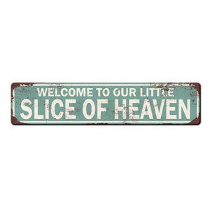 May include: Rectangular metal sign with a distressed teal background and white text that reads "WELCOME TO OUR LITTLE SLICE OF HEAVEN." The sign has a rusty brown border, giving it a vintage look.