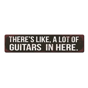 May include: A rustic brown metal sign with white text that reads "THERE'S LIKE, A LOT OF GUITARS IN HERE."