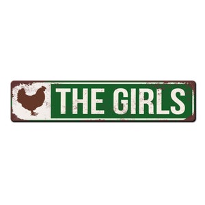 Puede incluir: Un letrero de metal oxidado con fondo verde y texto blanco que dice "THE GIRLS". Una silueta marr&oacute;n de una gallina est&aacute; en el lado izquierdo del letrero.