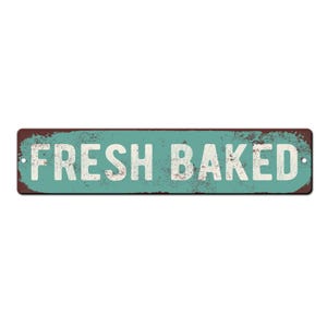 May include: A rectangular metal sign with the words "FRESH BAKED" in white block letters. The sign has a distressed teal background and a rusty brown border, giving it a vintage look.