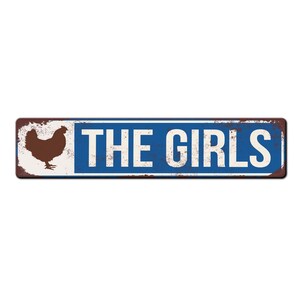 Puede incluir: Un letrero de metal azul y blanco con textura oxidada. El letrero tiene una silueta de un pollo en el lado izquierdo y el texto "THE GIRLS" en letras blancas en el lado derecho.