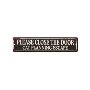 May include: A rectangular metal sign with the text "PLEASE CLOSE THE DOOR CAT PLANNING ESCAPE" in white letters on a dark brown background. The sign has a distressed, vintage look.