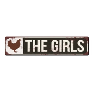 Puede incluir: Un letrero de metal oxidado con fondo blanco y texto negro que dice "THE GIRLS". Una silueta marr&oacute;n de una gallina est&aacute; en el lado izquierdo del letrero.