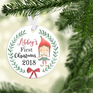 May include: White round Christmas ornament with a green wreath, a girl elf illustration, and the text "Abby's First Christmas 2018".