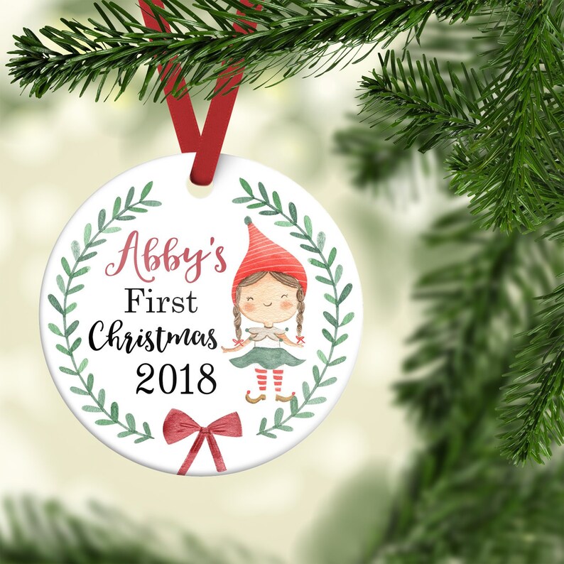 May include: White ceramic Christmas ornament with a green wreath, a cartoon elf girl, and the text "Abby's First Christmas 2018".