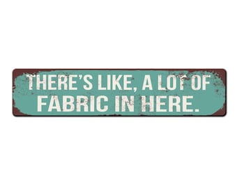 May include: A teal blue and brown rectangular sign with the text "THERE'S LIKE, A LOT OF FABRIC IN HERE." in white lettering.