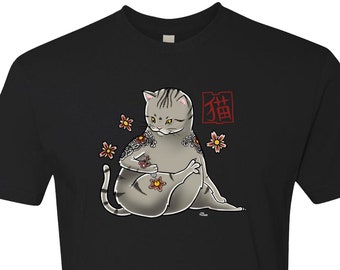Puede incluir: Camiseta negra con un gráfico de un gato gris con tatuajes rojos y amarillos. El gato está sentado con sus patas metidas debajo de su cuerpo. La palabra japonesa para "gato" está escrita en rojo sobre el gato.