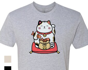 Puede incluir: Una camiseta gris con una imagen de dibujos animados de un Maneki-Neko, un gato de la suerte japonés, blanco y gris, que sostiene una bolsa de monedas de oro y un pez. El gato está sentado en un cojín rojo con ribetes dorados.