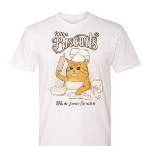 May include: White t-shirt with a cartoon cat wearing a chef's hat and making biscuits. The text on the shirt reads "Kitty's Biscuits Made from Scratch".