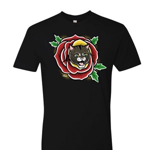 May include: Black t-shirt featuring a colorful graphic of a cat's face inside a red rose. The cat has yellow eyes and is surrounded by green leaves. The design is reminiscent of traditional tattoo art.