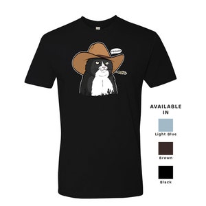 May include: Black and white cat wearing a brown cowboy hat with the word "MEOWDY" above it. The cat is wearing a black shirt with a white collar. The shirt is available in light blue, brown, and black.