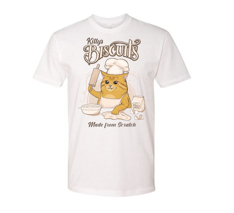 May include: White t-shirt with a cartoon cat wearing a chef's hat and making biscuits. The text on the shirt reads "Kitty's Biscuits Made from Scratch".