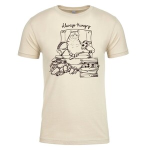 May include: A beige t-shirt with a black and white illustration of a cat sitting in a chair with pizza and other food around it. The text "Always Hungry" is above the cat.