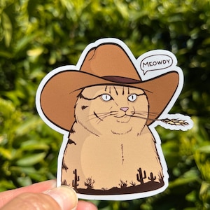 May include: A cartoon cat wearing a brown cowboy hat with the text "MEOWDY" in a speech bubble. The cat is holding a stalk of wheat in its mouth and is standing in front of a desert landscape with cacti.