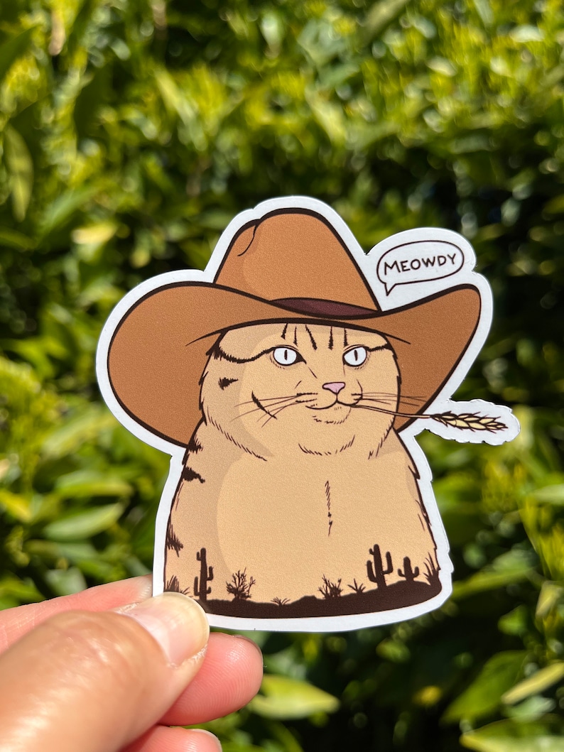 May include: A cartoon cat wearing a brown cowboy hat with the text "MEOWDY" in a speech bubble. The cat is holding a stalk of wheat in its mouth and is standing in front of a desert landscape with cacti.