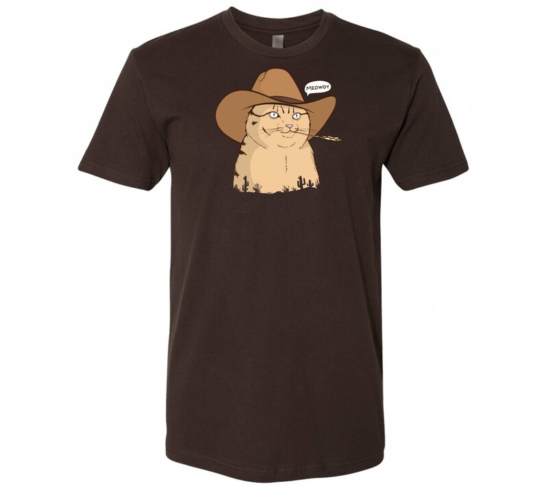 May include: A brown t-shirt with a cartoon image of a cat wearing a cowboy hat and holding a piece of grass in its mouth. The cat is looking to the right and has a speech bubble above its head that says "MEOWDY".