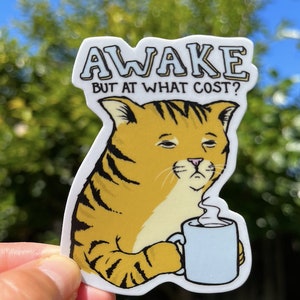 May include: A sticker featuring a cartoon cat with a tired expression, holding a blue mug with steam coming from it. The text "AWAKE BUT AT WHAT COST?" is above the cat.