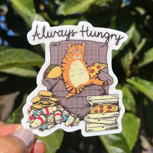 May include: A sticker featuring a cartoon orange cat sitting in a chair with a slice of pizza and a taco. The cat is surrounded by empty food containers and the text "Always Hungry" is at the top of the sticker.