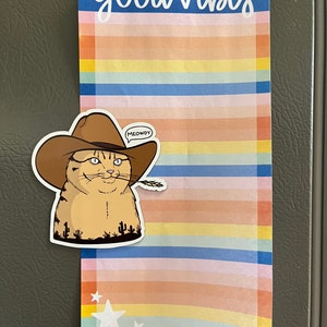 May include: A colorful striped notepad with the text "good vibes" at the top. A sticker of a cat wearing a cowboy hat and holding a piece of wheat is attached to the notepad. The sticker has a speech bubble that says "MEOWDY".
