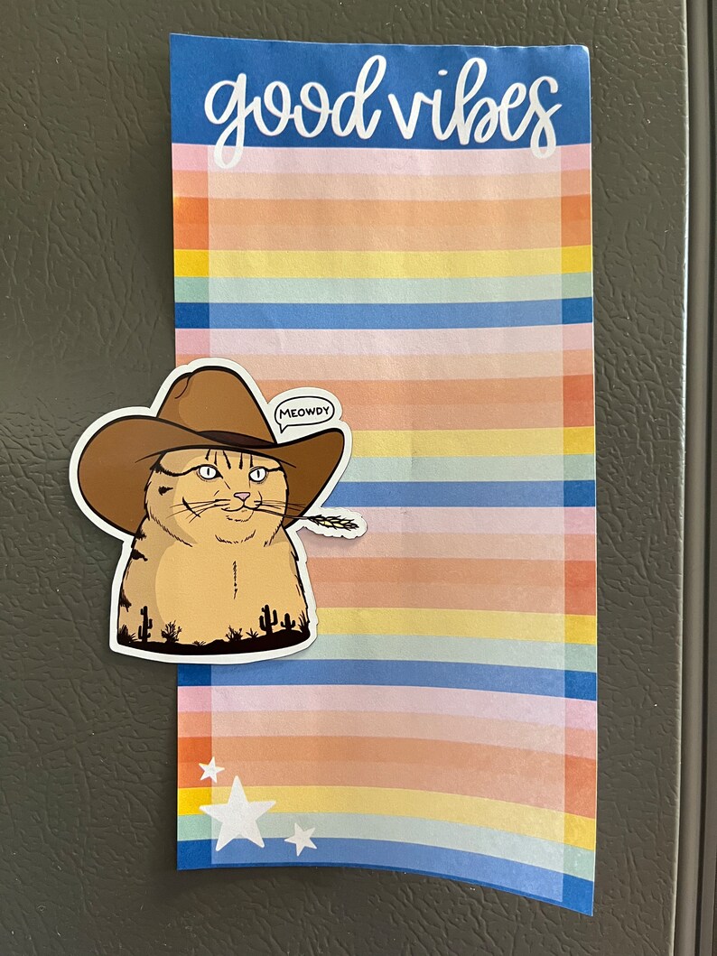 May include: A colorful striped notepad with the text "good vibes" at the top. A sticker of a cat wearing a cowboy hat and holding a piece of wheat is attached to the notepad. The sticker has a speech bubble that says "MEOWDY".