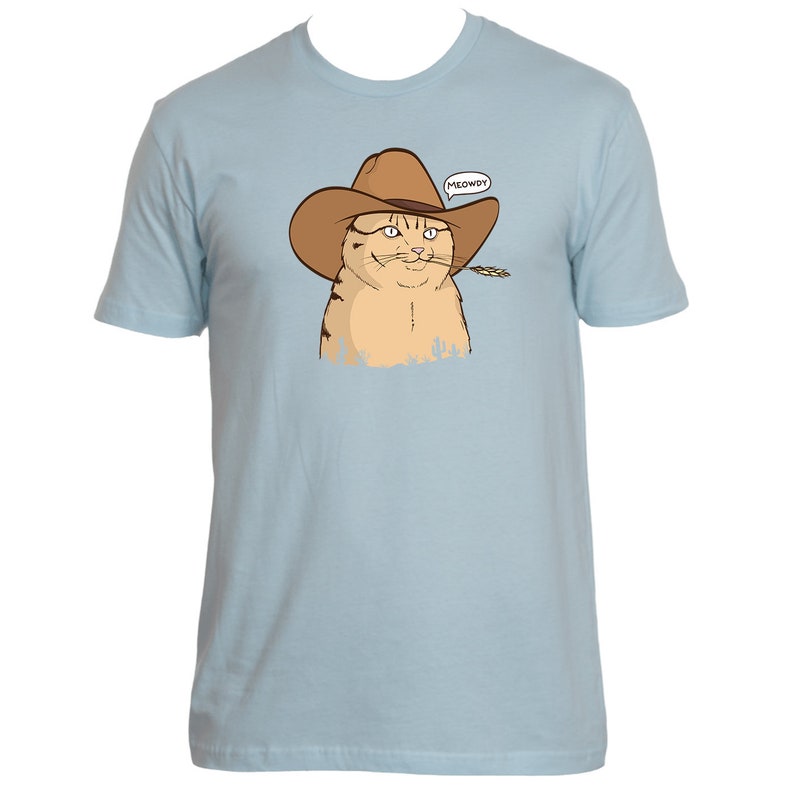 May include: A light blue t-shirt with a cartoon image of a cat wearing a cowboy hat and holding a piece of grass in its mouth. The cat is looking to the right and has a speech bubble above its head that says "MEOWDY!"