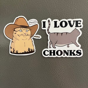 May include: Two cat-themed refrigerator magnets. The first magnet features a cartoon cat wearing a cowboy hat with the text "MEOWDY" in a speech bubble. The second magnet features a cartoon cat walking with the text "I LOVE CHONKS".