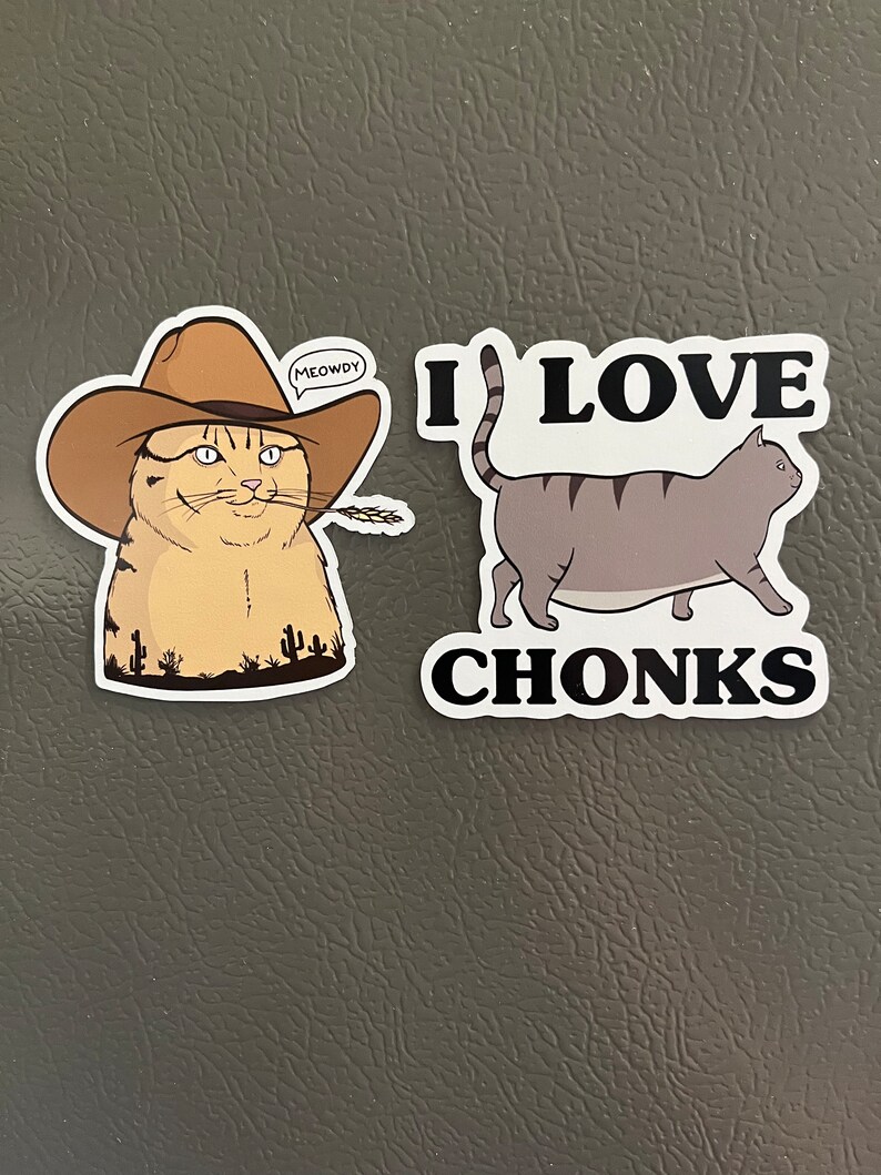 May include: Two cat-themed refrigerator magnets. The first magnet features a cartoon cat wearing a cowboy hat with the text "MEOWDY" in a speech bubble. The second magnet features a cartoon cat walking with the text "I LOVE CHONKS".