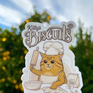 May include: A sticker featuring a cartoon cat chef with the text "Kitty's Biscuits Made from Scratch." The cat is wearing a chef's hat and apron, holding a rolling pin and working with dough. A bag of flour and bowl are also shown.
