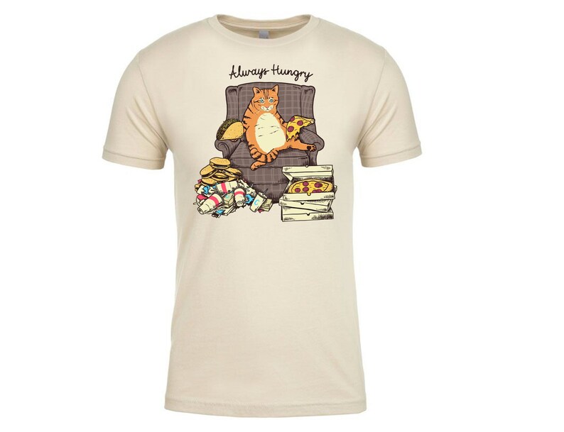 May include: A beige t-shirt with a cartoon illustration of a cat sitting in a chair and eating pizza. The text "Always Hungry" is above the cat.