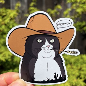 May include: A black and white cat wearing a brown cowboy hat with the word "MEOWDY" in a speech bubble. The cat is holding a stalk of wheat in its mouth and is standing in front of a desert landscape with cacti.