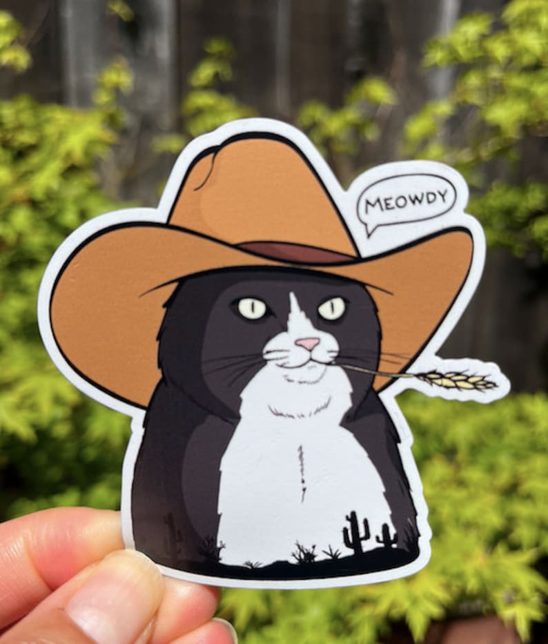 May include: A black and white cat wearing a brown cowboy hat with the word "MEOWDY" in a speech bubble. The cat is holding a stalk of wheat in its mouth and is standing in front of a desert landscape with cacti.