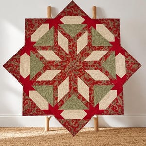 May include: A star-shaped quilt featuring a geometric pattern with red fabric displaying a dark paisley print, light beige diamond shapes, and muted green triangular sections. The textile piece is constructed with intricate stitching.