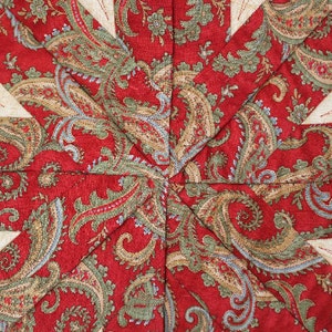 Puede incluir: Primer plano de un textil acolchado con un dise&ntilde;o de estrella. El patr&oacute;n central presenta una tela de cachemira roja, con acentos de verde, azul y dorado. Las puntas de la estrella son de color crema con detalles de hojas bordadas.