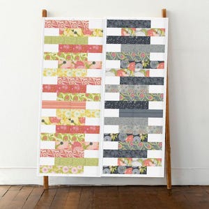 Könnte beinhalten: Eine Steppdecke mit Patchwork-Muster in zwei verschiedenen Farbschemata. Eine Seite ist eine Mischung aus rosa, gelben und grünen Blumenmustern, während die andere Seite eine Kombination aus grauen und schwarzen Paisley-Mustern ist. Die Steppdecke wird auf einem Holzständer vor einer weißen Wand präsentiert.