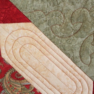 Puede incluir: Primer plano de una tela acolchada con una combinaci&oacute;n de colores beige, rojo y verde. El dise&ntilde;o presenta una forma alargada beige con costuras rojas, rodeada de estampados de cachemira rojos y florales verdes. El acolchado es detallado.
