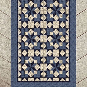 May include: A rectangular quilt featuring a repeating pattern of blue and cream-coloured star and diamond shapes. The quilt has a blue border and is displayed on a light-coloured surface.