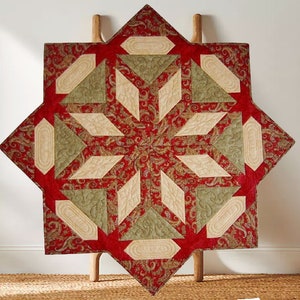 Può includere: Un runner trapuntato a forma di stella con un motivo paisley rosso. Il design include forme geometriche beige e verde oliva. Il runner è esposto su una scala di legno, mostrando il suo fascino decorativo.