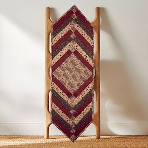 May include: A decorative table runner with a diamond-shaped design. The runner features alternating bands of burgundy, tan, and patterned fabric with floral and paisley motifs. It is displayed on a wooden ladder-style rack.
