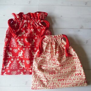 May include: Two fabric drawstring gift bags with red braided cords. The larger bag is red with a white pattern of kangaroos, hearts, and trees. The smaller bag is cream with red text reading Merry Christmas, Peace on Earth, and Joy to the World.