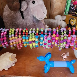 May include: A collection of colorful beaded bracelets displayed on a black horizontal bar. The bracelets feature various round beads in shades of pink, purple, yellow, green, and blue, with some including star-shaped accents and textured surfaces.