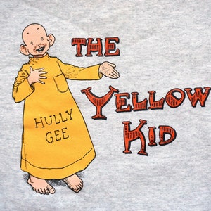May include: A gray t-shirt with a cartoon illustration of a bald, yellow character with large ears and a yellow robe. The text "The Yellow Kid" is printed in red letters on the shirt. The character is holding a hand up in a gesture of greeting. The text "Hully Gee" is printed on the character's robe.