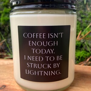 May include: A glass jar candle with a black lid and a black label. The label reads "COFFEE ISN'T ENOUGH TODAY. I NEED TO BE STRUCK BY LIGHTNING." The candle has a creamy white wax and is set on a wooden surface.