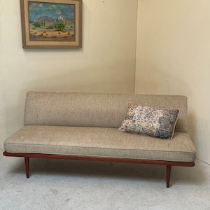 May include: A light brown fabric daybed with a dark wood frame and legs. A patterned pillow is on the daybed. A framed painting of a desert landscape hangs on the wall behind the daybed.