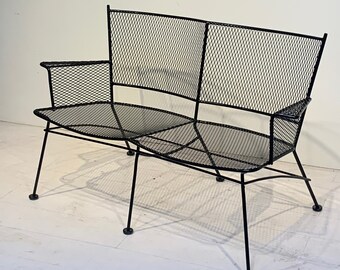 May include: Black metal outdoor bench with a mesh seat and back. The bench has a two-seat design with armrests and a minimalist, geometric style. The legs are thin and angled, with small round feet.