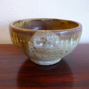 May include: A handcrafted ceramic bowl with a rustic aesthetic. The bowl features a rounded shape with a wide opening and a textured surface. The exterior showcases a blend of brown, tan, and cream colors, with visible text. The bowl sits on a dark wooden surface.