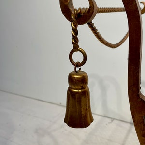 Puede incluir: Primer plano de un objeto decorativo de metal dorado y ornamentado. El objeto presenta un colgante en forma de campana que cuelga de una cuerda trenzada y un anillo. El dise&ntilde;o incluye elementos curvos y enrollados, lo que sugiere un estilo vintage o antiguo.