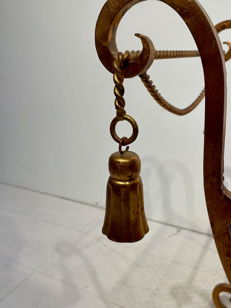 Puede incluir: Primer plano de un objeto decorativo de metal dorado y ornamentado. El objeto presenta un colgante en forma de campana que cuelga de una cuerda trenzada y un anillo. El dise&ntilde;o incluye elementos curvos y enrollados, lo que sugiere un estilo vintage o antiguo.